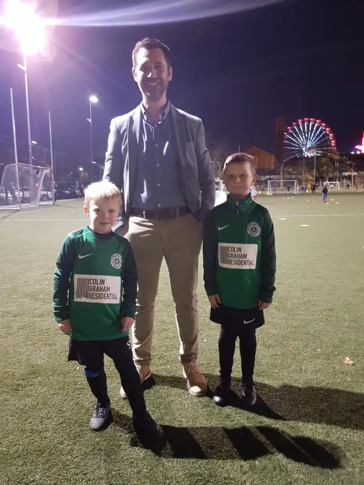 A member of the Colin Graham Residential team with two young footballers in green kit sponsored by the company, on a floodlit pitch